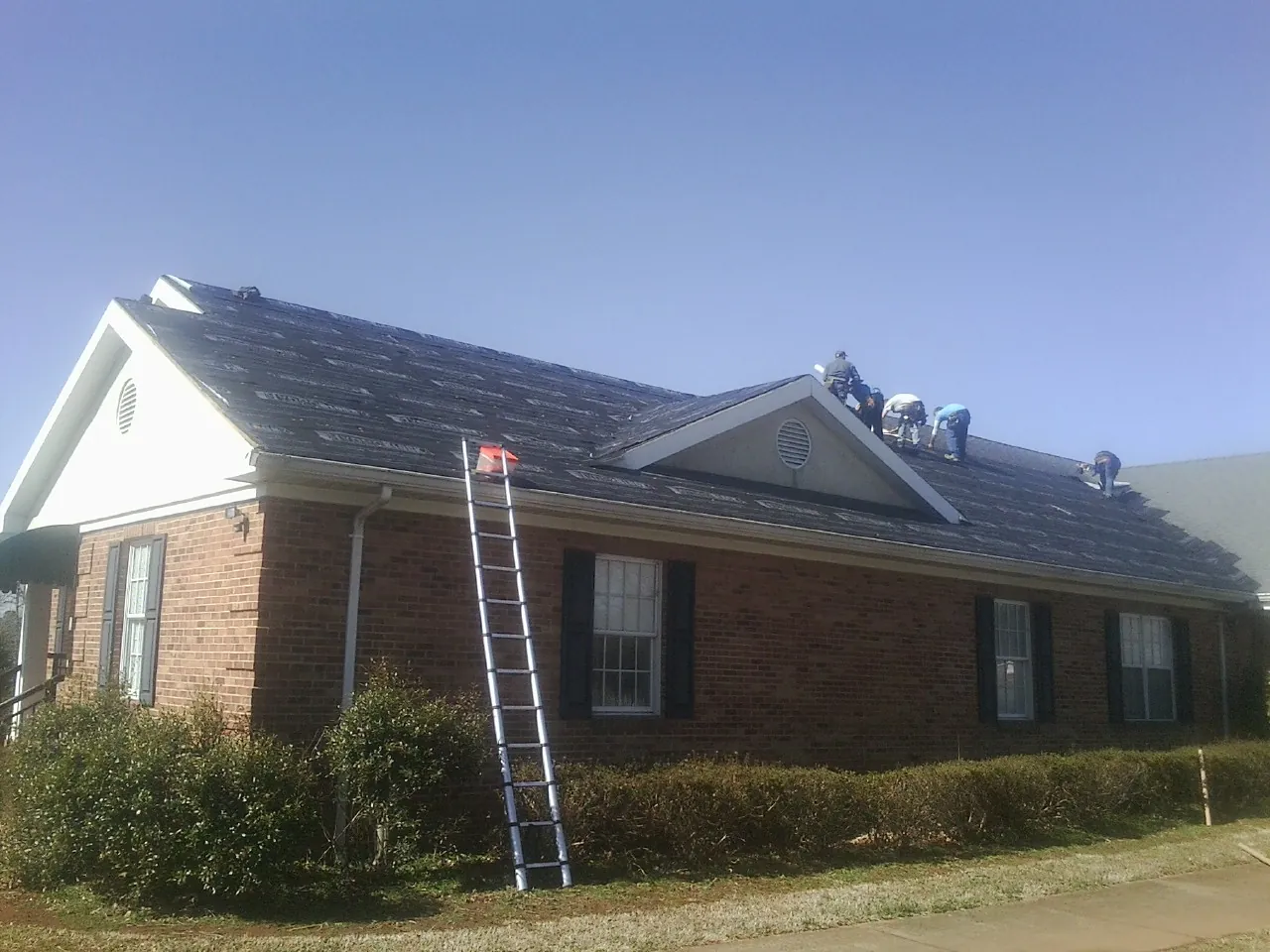 Crew tearing shingles off a brick ranch roof with a ladder set — Metal Roof Repair work by our crew in Colchester